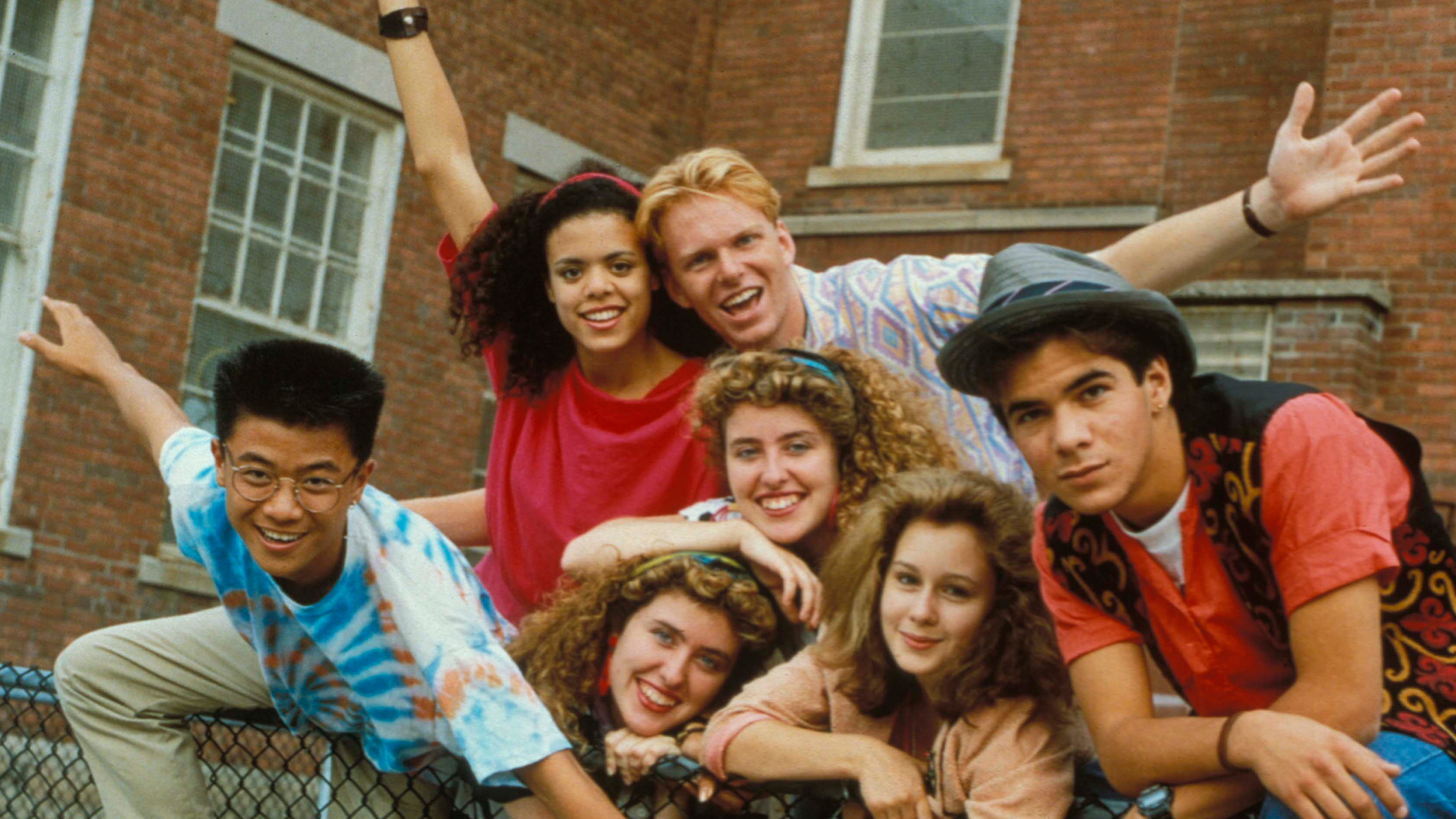 A group of teenagers from the 1990s leaning on a fence and posing for a photo in front of a high school. 