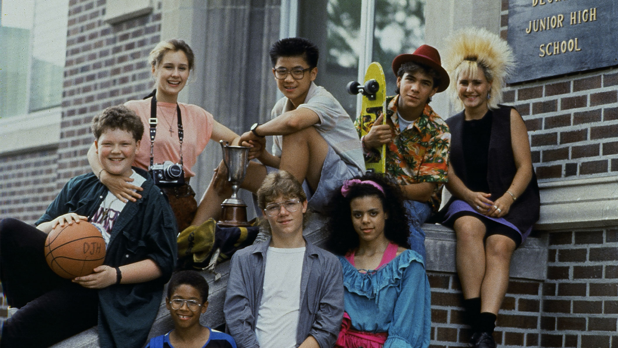 A group of teenagers from the 1980s posing for a photo in front of a high school. 